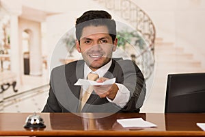 Happy smiling receptionist in hotel giving key to guest and papers to sign, hotel background