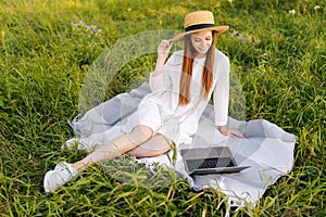 Happy smiling red-haired young woman wearing straw hat and white dress looking to laptop screen sitting on beautiful