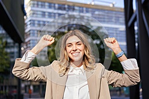 Happy smiling saleswoman in beige suit, celebrates her achievement, triumphing, making fist pump and looking excited on