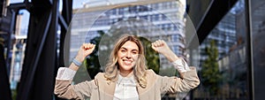 Happy smiling saleswoman in beige suit, celebrates her achievement, triumphing, making fist pump and looking excited on
