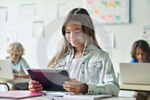 Happy smiling schoolgirl reading task from tablet computer device in classroom.