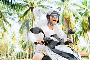 Happy smiling and screaming male tourist in helmet and sunglasses riding motorbike scooter during his tropical vacation under palm