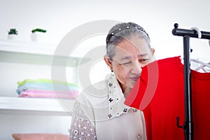 Happy smiling senior elderly woman housewife hanging clothes indoor to dry, sniffing from clothes, older Asian female doing