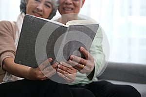 Happy smiling senior family couple in love reading book together, hugging embracing while spending free time at home on retirement