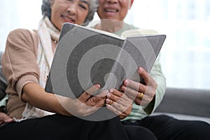 Happy smiling senior family couple in love reading book together, hugging embracing while spending free time at home on retirement