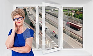 Happy smiling senior woman stands inside near threefold pvc window pane with hoisy highway with cars on background