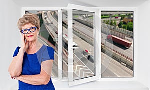 Happy smiling senior woman stands inside near threefold pvc window pane with hoisy highway with cars on background photo