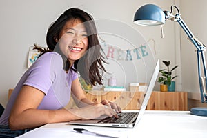 Happy, smiling teen asian female college student studying at home, doing homework using laptop looking at camera.