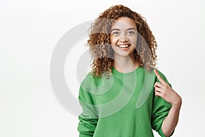 Happy smiling teenage girl, curly-female model pointing fingers at herself, self-promoting, standing over white
