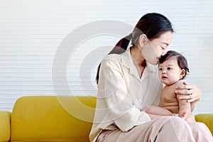 Happy smiling toddle baby sitting on mother lap on yellow sofa in living room at home, mom kissing her little girl daughter, photo