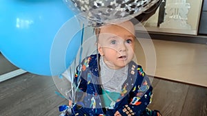 Happy smiling toddler boy sitting on floor and playing with colorful air balloons on his first birthday party