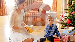 Happy smiling toddler boy sitting under Christmas tree with mother and holding gifts in boxes