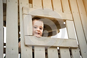 A happy smiling toddler boy of two or three years looks out of a window on a wooden playground. Toddlerhood and childhood concept