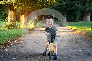 A happy smiling toddler boy of two or three years old rides a bicycle or balance bike in a city park on a sunny summer day.