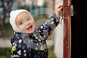 Happy smiling toddler in white woolen hat opening old door to somewhere. Children safety concept