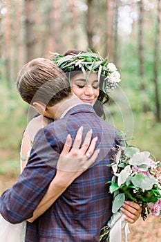 Happy and smiling wedding couple in the forest. Handsome groom embacing his pretty bride with floral wreath on the head