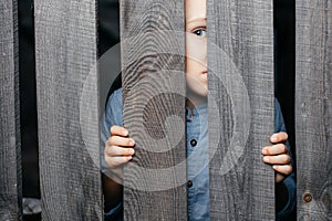 Happy smiling white boy looks out of the crack of a wooden fence. Childish curiosity. Espionage. Rural life. Child