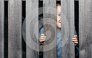 Happy smiling white boy looks out of the crack of a wooden fence. Childish curiosity. Espionage. Rural life. Child