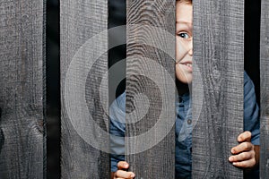 Happy smiling white boy looks out of the crack of a wooden fence. Childish curiosity. Espionage. Rural life. Child