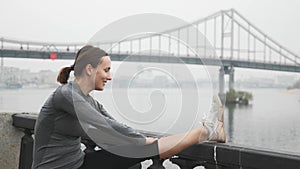 Happy smiling woman in activewear is stretching on city embankment with concrete bridge on background. Young female athlete stretc