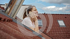 Happy, smiling woman as she looks out of an open attic window, engaged in a phone conversation, and admires the red