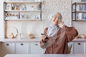 Happy and smiling woman dancing and singing at home in kitchen, Muslim woman in hijab and headphones listening to music