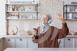 Happy and smiling woman dancing and singing at home in kitchen, Muslim woman in hijab and headphones listening to music