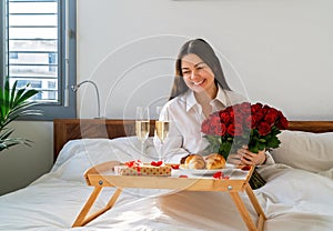 Happy smiling woman holding bouquet of flowers having surprise gift and romantic breakfast in bed on Valentine Day