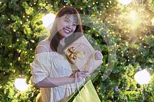 Happy smiling woman holding a golden gift box and shopping bag with lights on Christmas tree background. Happy