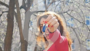 Happy smiling woman holding her new apartment or car keys in one hand with red manicure outdoors during spring time on