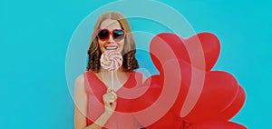 happy smiling woman holding lollipop and bunch of red heart shaped balloons wearing a sunglasses on a blue background