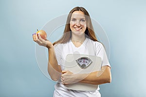 Happy smiling woman holding weight scales and red apple on a blue background. Healthy eating concept. Diet