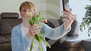 Happy smiling woman holds bunch of flowers and looks at smartphone display and receives messages of congratulation.