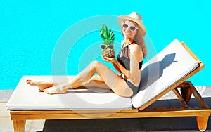 happy smiling woman lies on deckchair with pineapple over blue water pool background