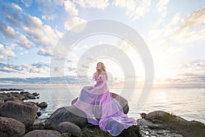 Happy smiling woman with long blonde hair in fashionable pink summer dress against sunset sky clouds and sea