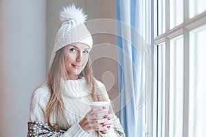 Happy smiling woman with long brown hair wearing season casual clothes white sweater and knitted hat holding hot dreank