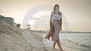 Happy smiling woman in long skirt walking on the sandy sea beach at sunset. Concept of happiness, travel, journey, trip.