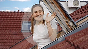 Happy smiling woman looking out of the attic window and waving hand in camera