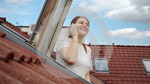 Happy smiling woman looking out of the open attic window and talking by phone while enjoying view on red tiled roof of