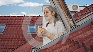 Happy smiling woman looks through the open attic window and drinks tea or coffee from a mug in morning.