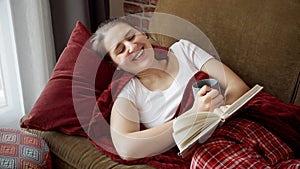 Happy smiling woman lying on sofa a book and laughing. People resting at home, hobby, education, female leisure