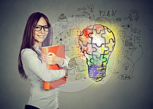 Happy smiling woman with orange folder notebook near a concrete wall with a bright puzzle light bulb business id