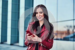 Happy smiling woman in red jacket is standing next to glass building and chating with someone by mobile phone