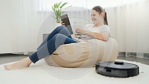 Happy smiling woman relaxing in chair with cup of tea and tablet computer while robot vacuum cleaner doing housework and