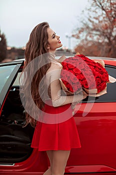 Happy smiling woman with rose bouquet of flowers posing by new red car. Beauty outdoor autumn portrait. Valentines day. Romantic