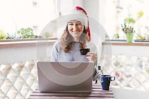 Happy smiling woman sitting at the table with laptop and credit card wearing Santa`s red hat