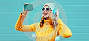 Happy smiling woman taking selfie picture by phone while listening to music in wireless headphones wearing yellow knitted sweater