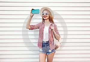 Happy smiling woman taking selfie picture by phone in summer round straw hat, checkered shirt, shorts on white wall