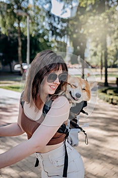 Happy smiling woman traveler is riding her electro scooter in city parkland with dog Welsh Corgi Pembroke in a special backpack