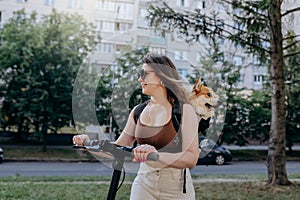 Happy smiling woman traveler is riding her electro scooter in city parkland with dog Welsh Corgi Pembroke in a special backpack